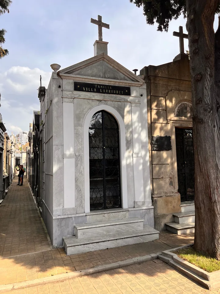 White marble family mausoleum topped by a cross, iron-and-glass door, on a narrow Recoleta lane