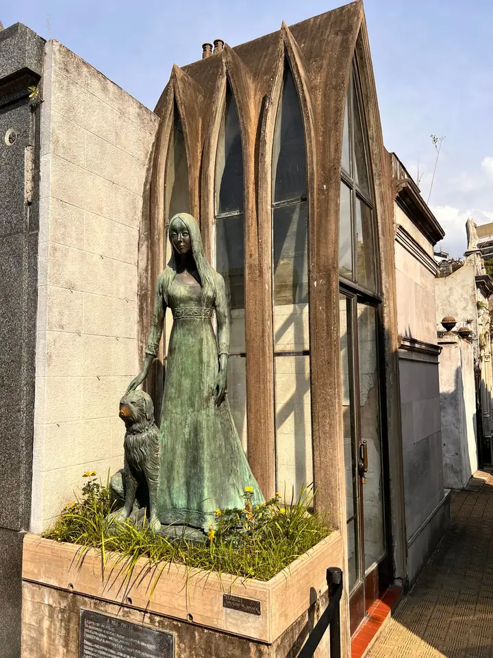 Green bronze statue of a young woman resting her hand on a dog, before a gothic glass chapel