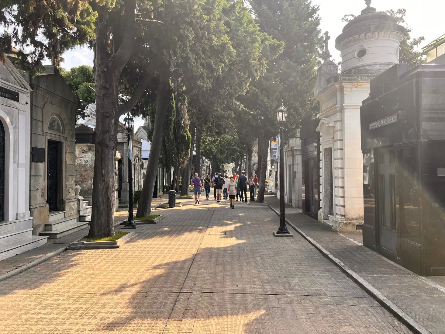 Wide tree-lined avenue of mausoleums and lamp posts, visitors strolling in warm late-afternoon light
