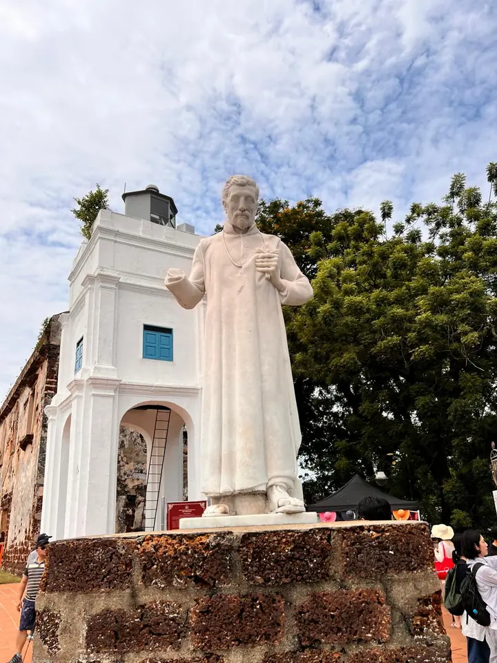 White marble statue of a robed missionary on a laterite plinth, whitewashed bell tower behind, Melaka